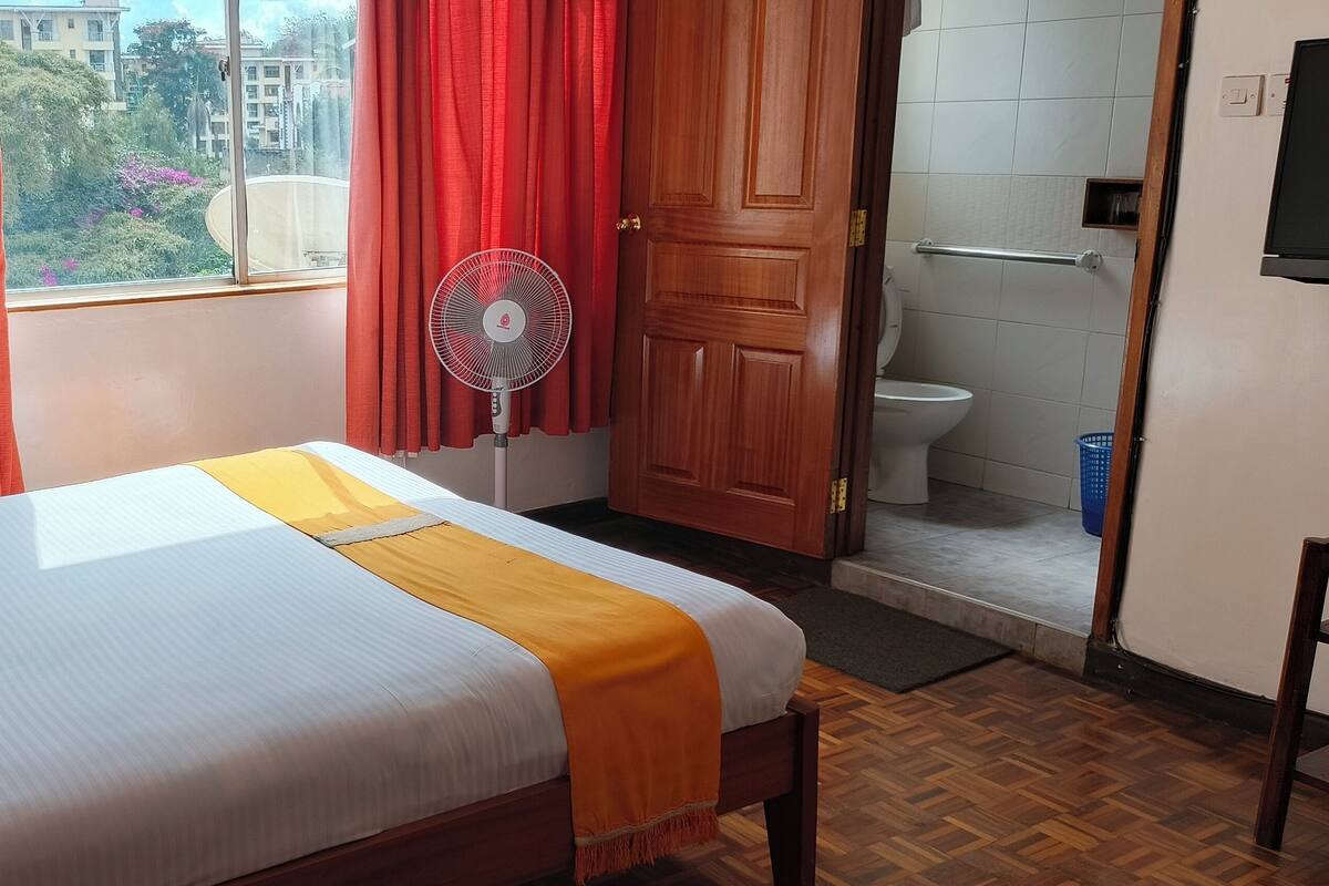 Habitación de hotel con cama, cortinas rojas, ventilador, puerta de madera y vistas a edificios.