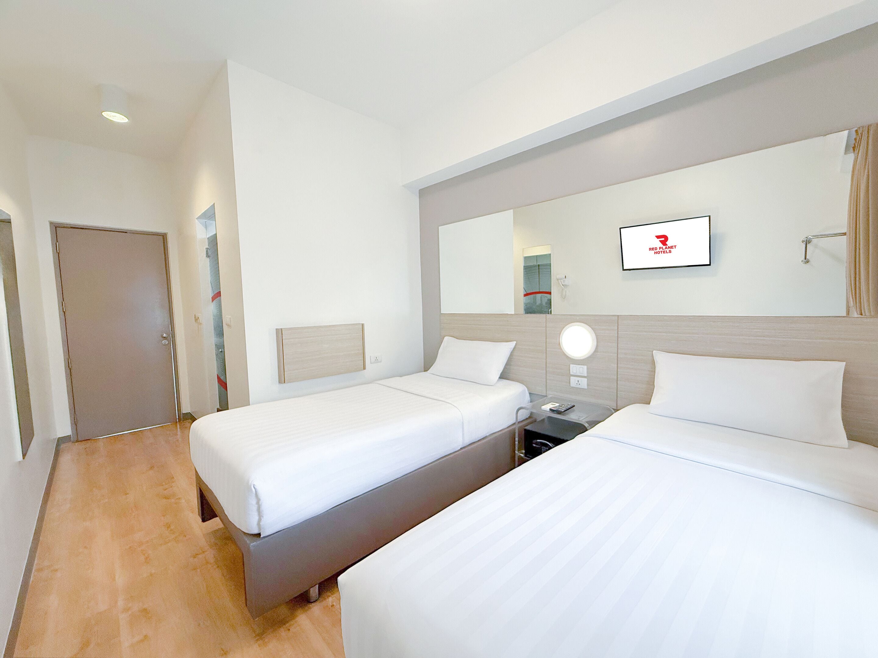 A hotel room with two single beds, a bedside table with a lamp, a mirror, and a wall-mounted TV.