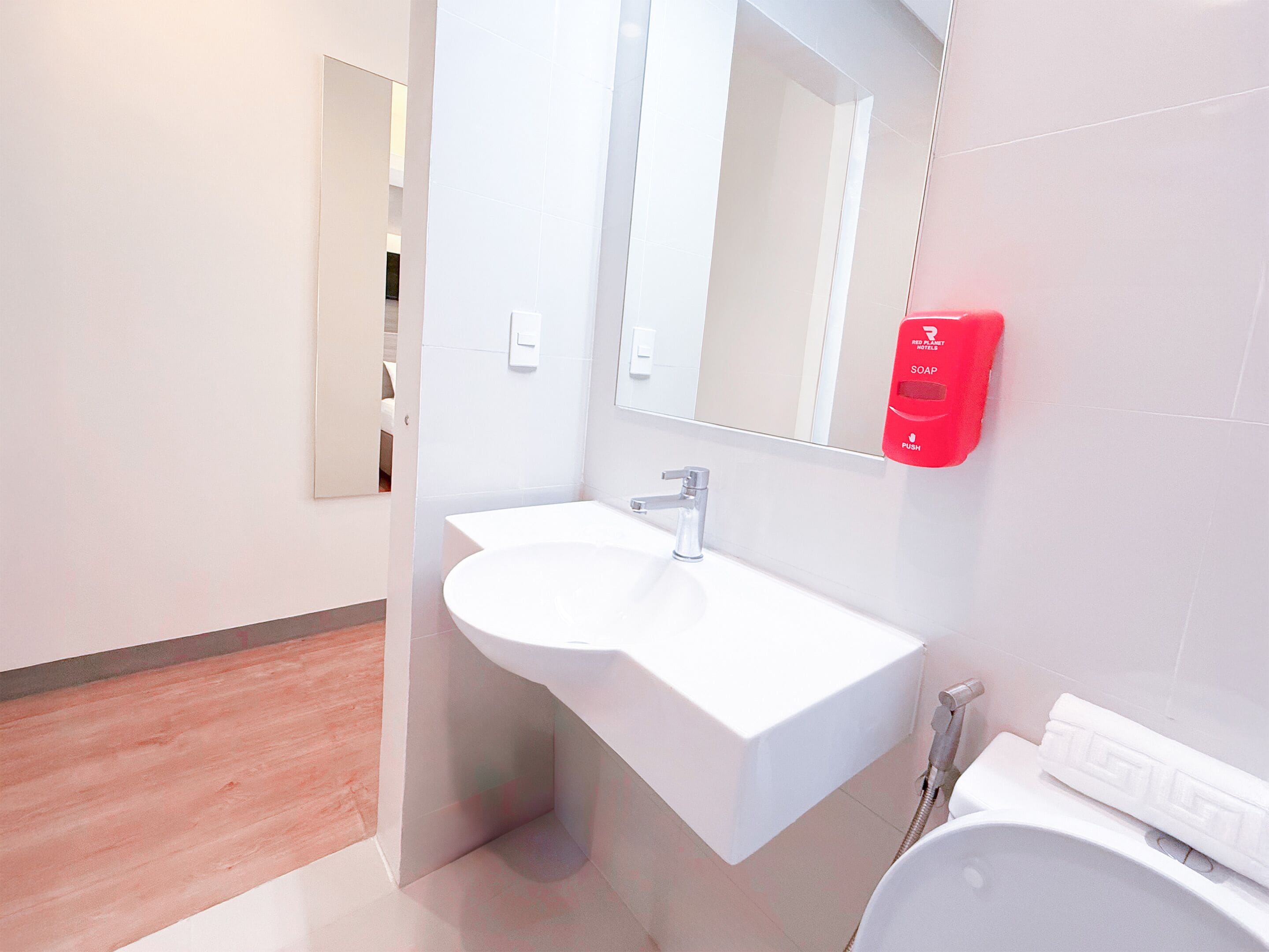 A modern bathroom with a white sink, a mirror, and a red soap dispenser.