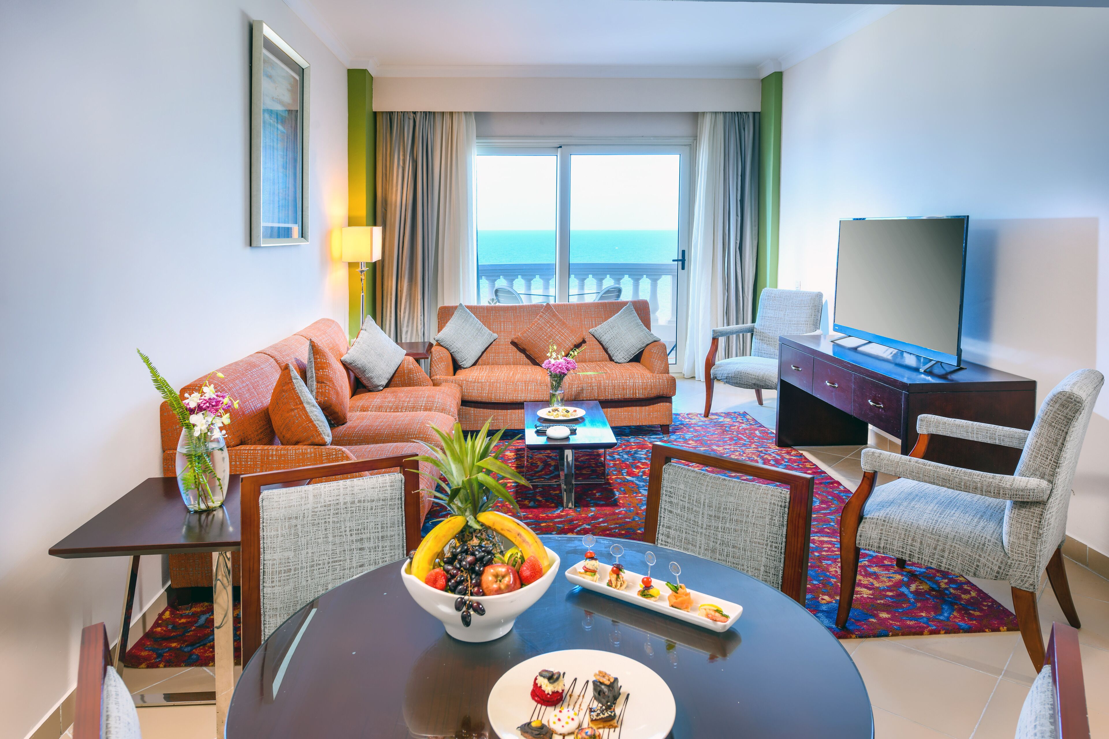 A hotel room with a sofa, dining table, chairs, a TV, and a balcony with a view of the sea.