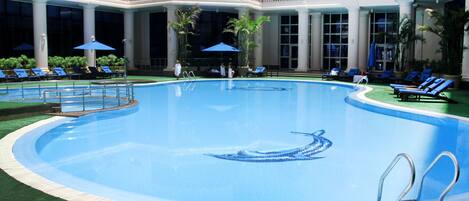 A large indoor swimming pool with a dolphin mosaic, surrounded by lounge chairs and umbrellas.