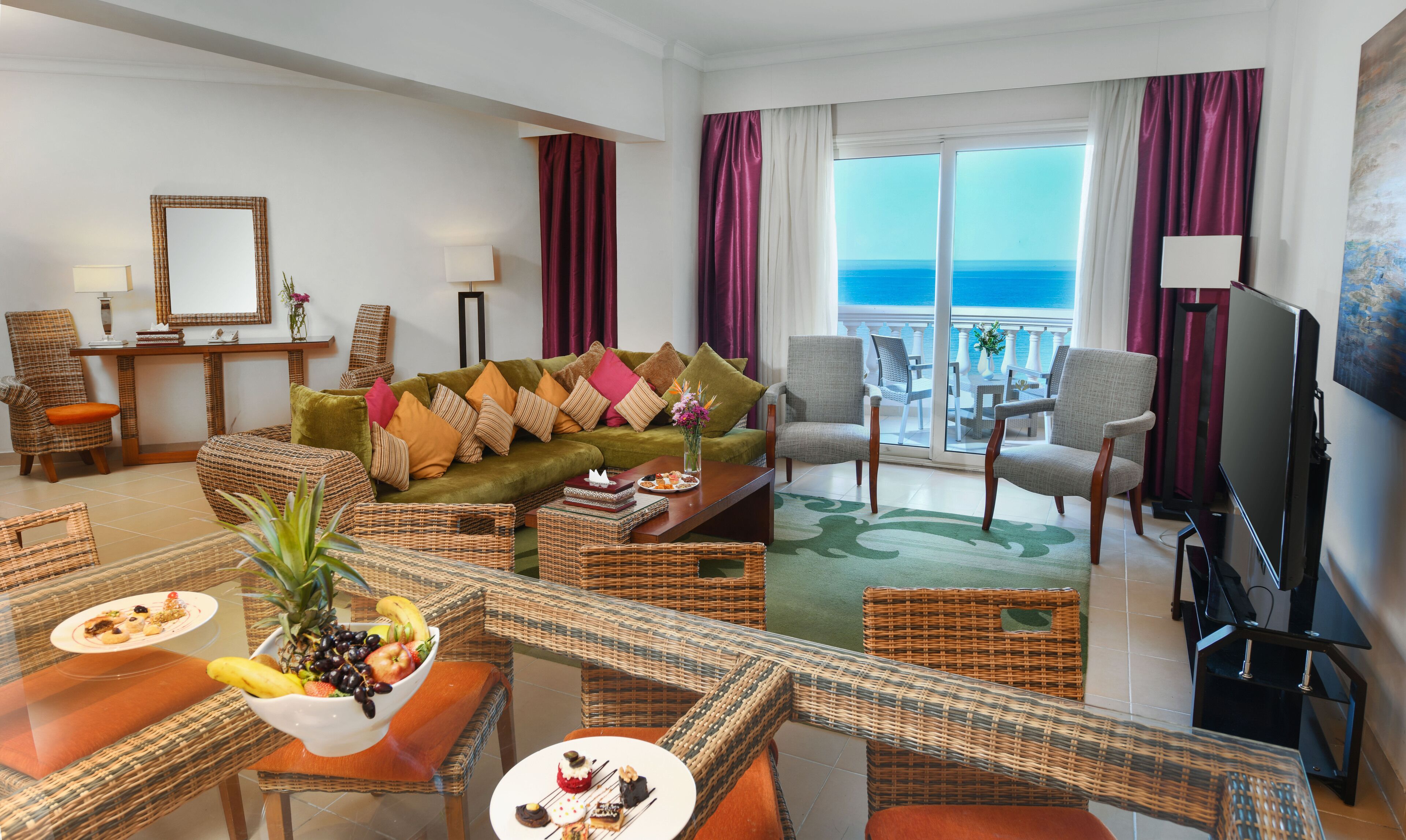 A hotel room with a green sofa, wicker furniture, a dining area with fruit, a TV, and a balcony with a view of the sea.