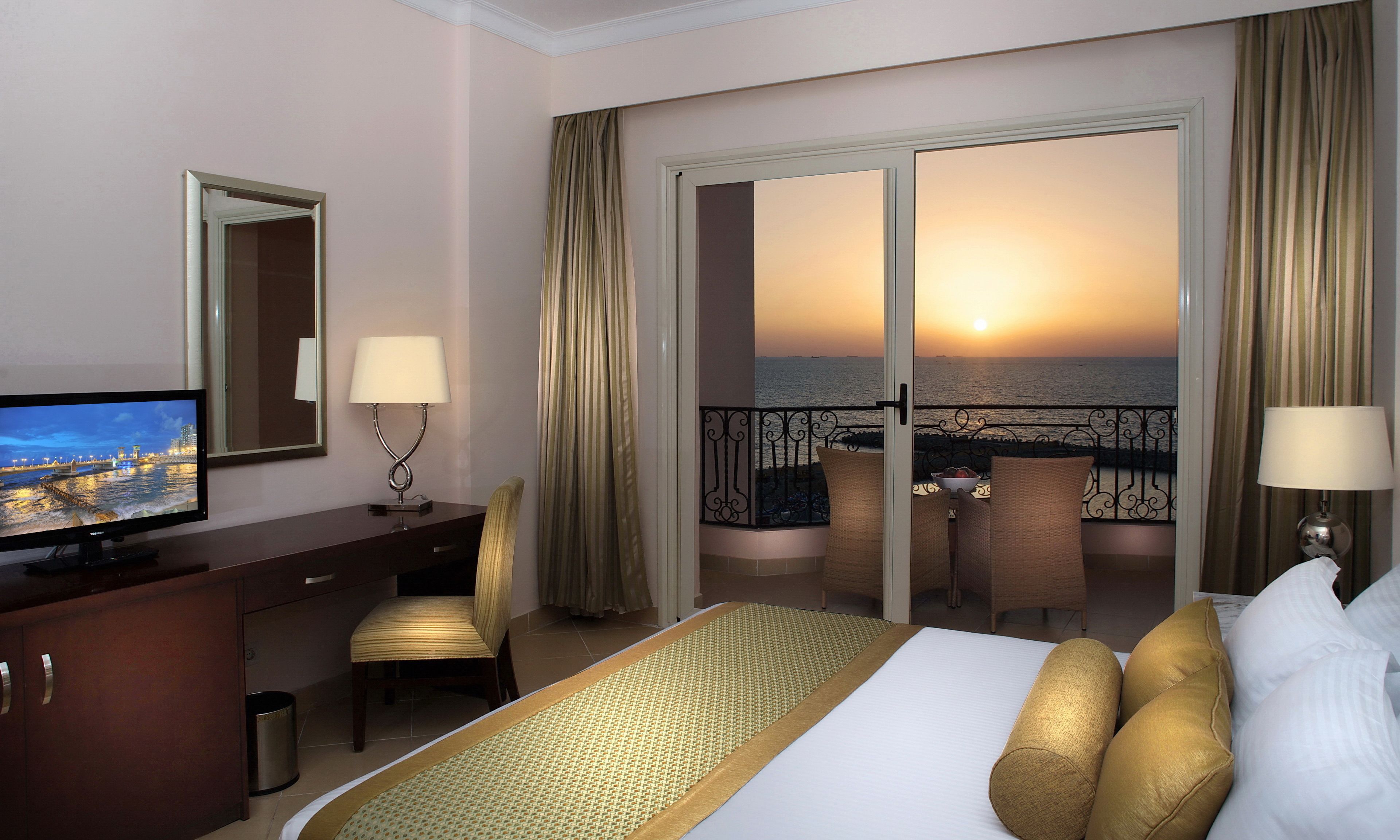 A hotel room with a bed, a desk with a TV, a balcony with a view of the sunset, and a chair.