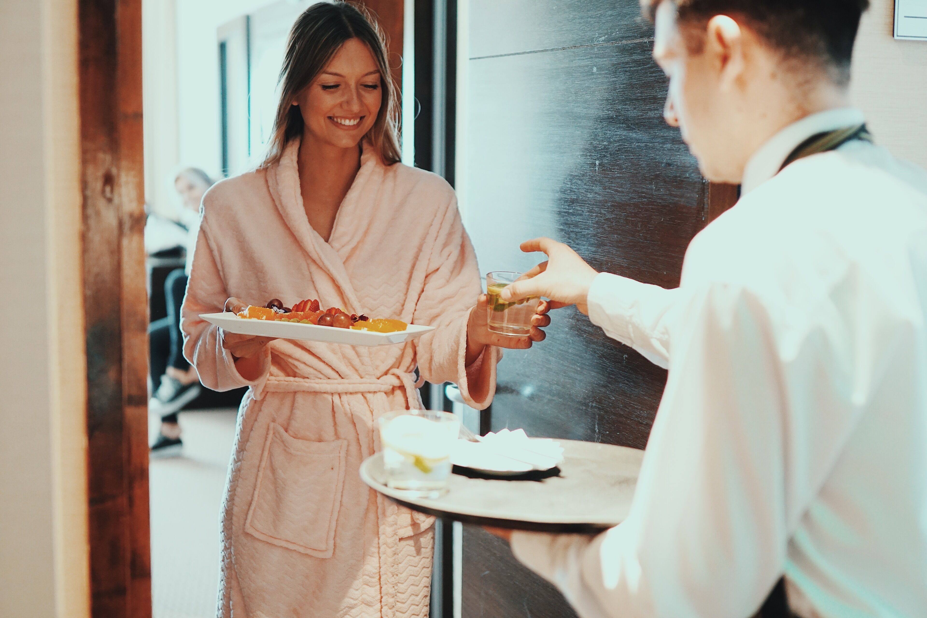 A person in a robe holding a tray with food and drinks, standing in a hallway with a wooden door and a glass wall.