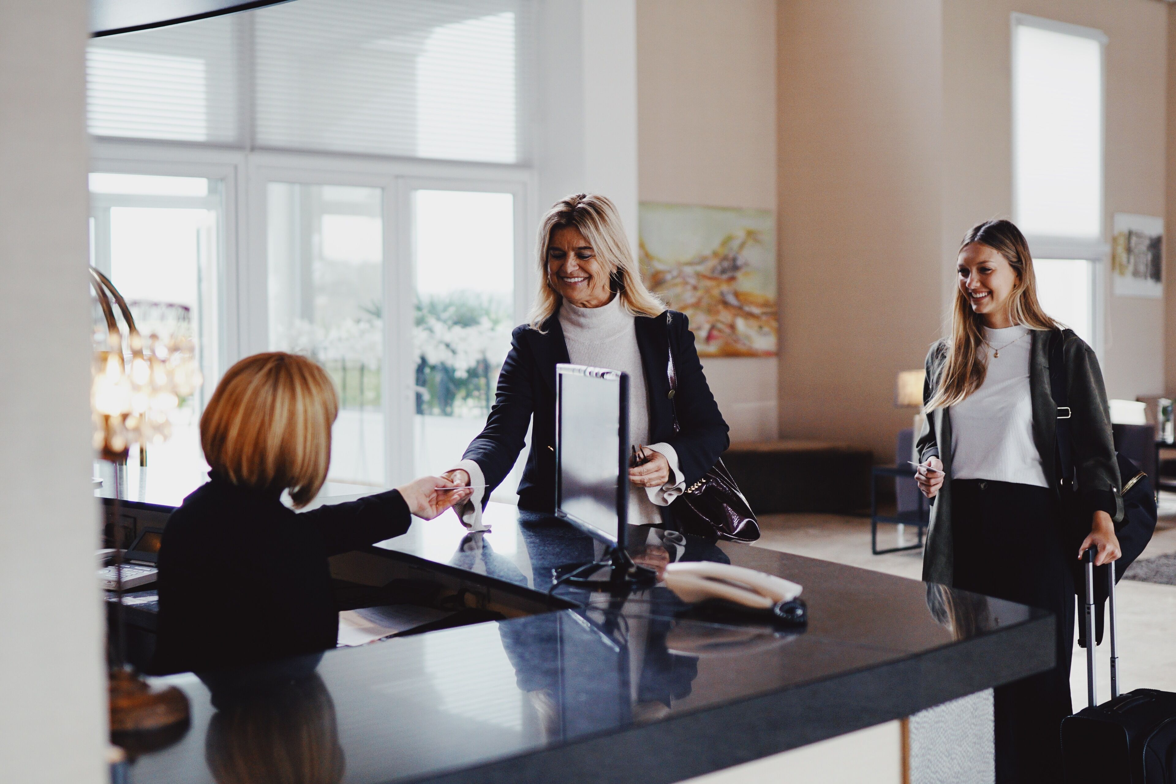 A reception desk in a hotel lobby with a glass partition, a large window, and a painting on the wall.
