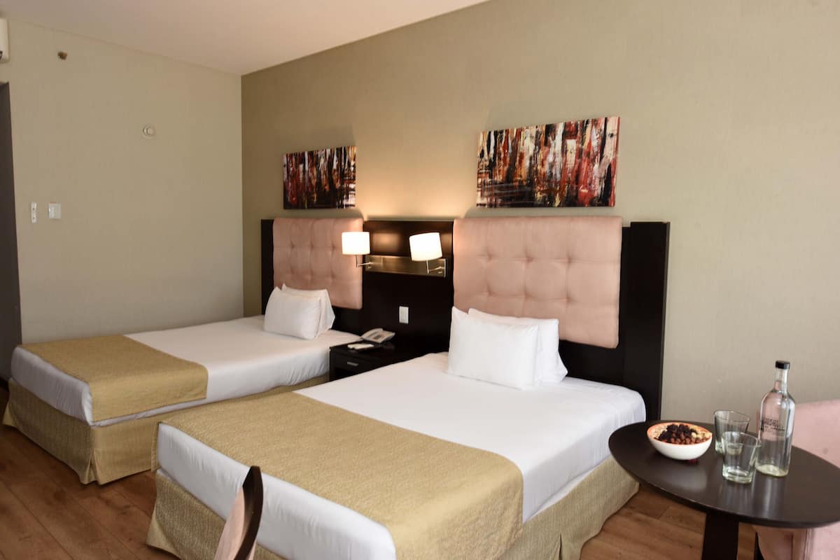 Ein Hotelzimmer mit zwei Betten, einem Nachttisch mit Lampe, einem kleinen Tisch mit Schüssel und Flasche sowie abstrakter Kunst an den Wänden.