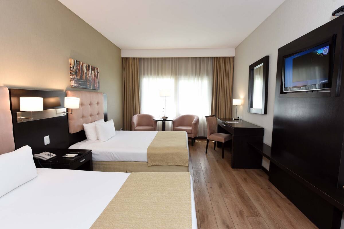 Ein Hotelzimmer mit zwei Betten, einem Schreibtisch, einem Sessel, einem Fernseher und einem Bild an der Wand.