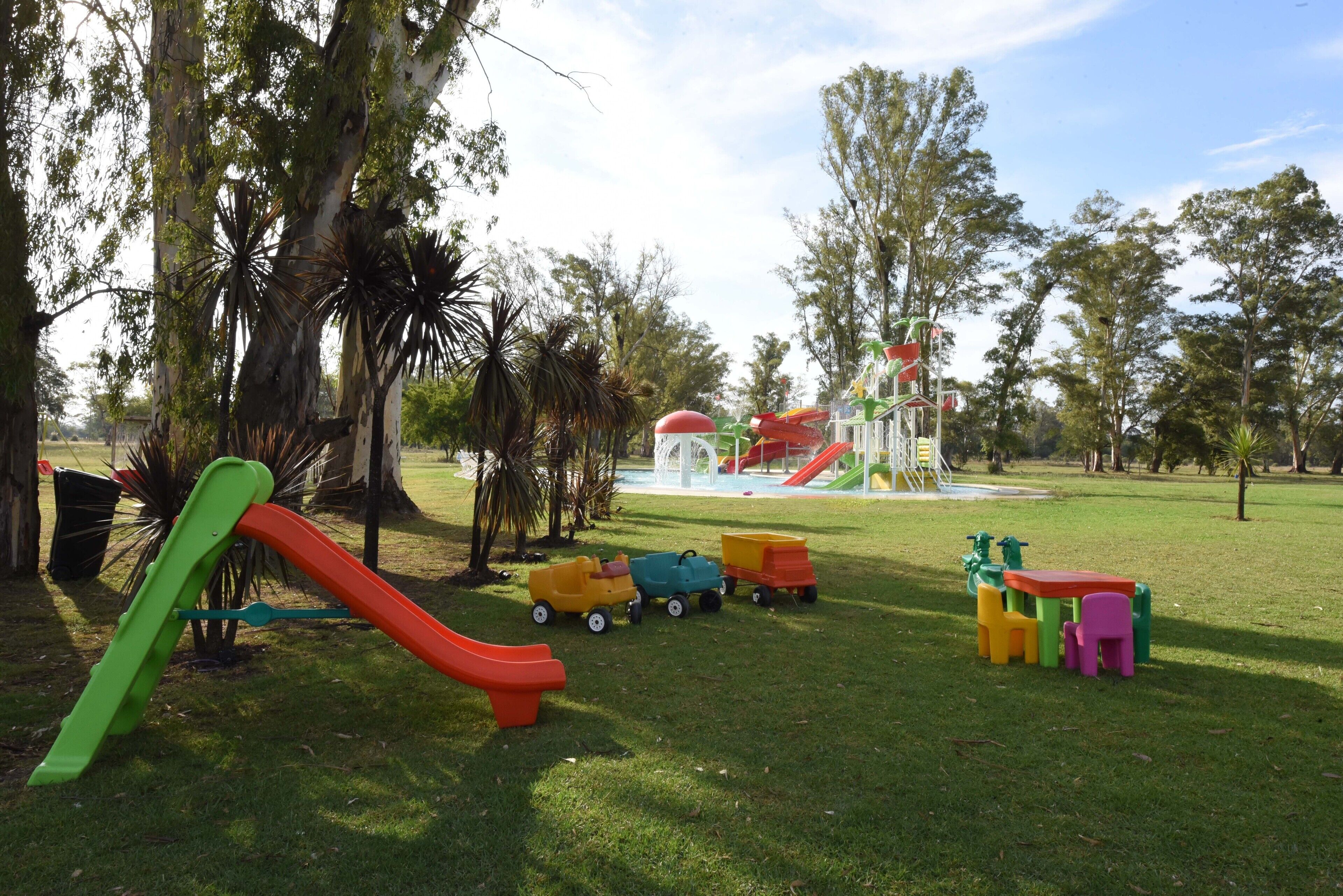 Un parque infantil con toboganes y camiones de juguete, rodeado de árboles y una zona de juegos acuáticos al fondo.