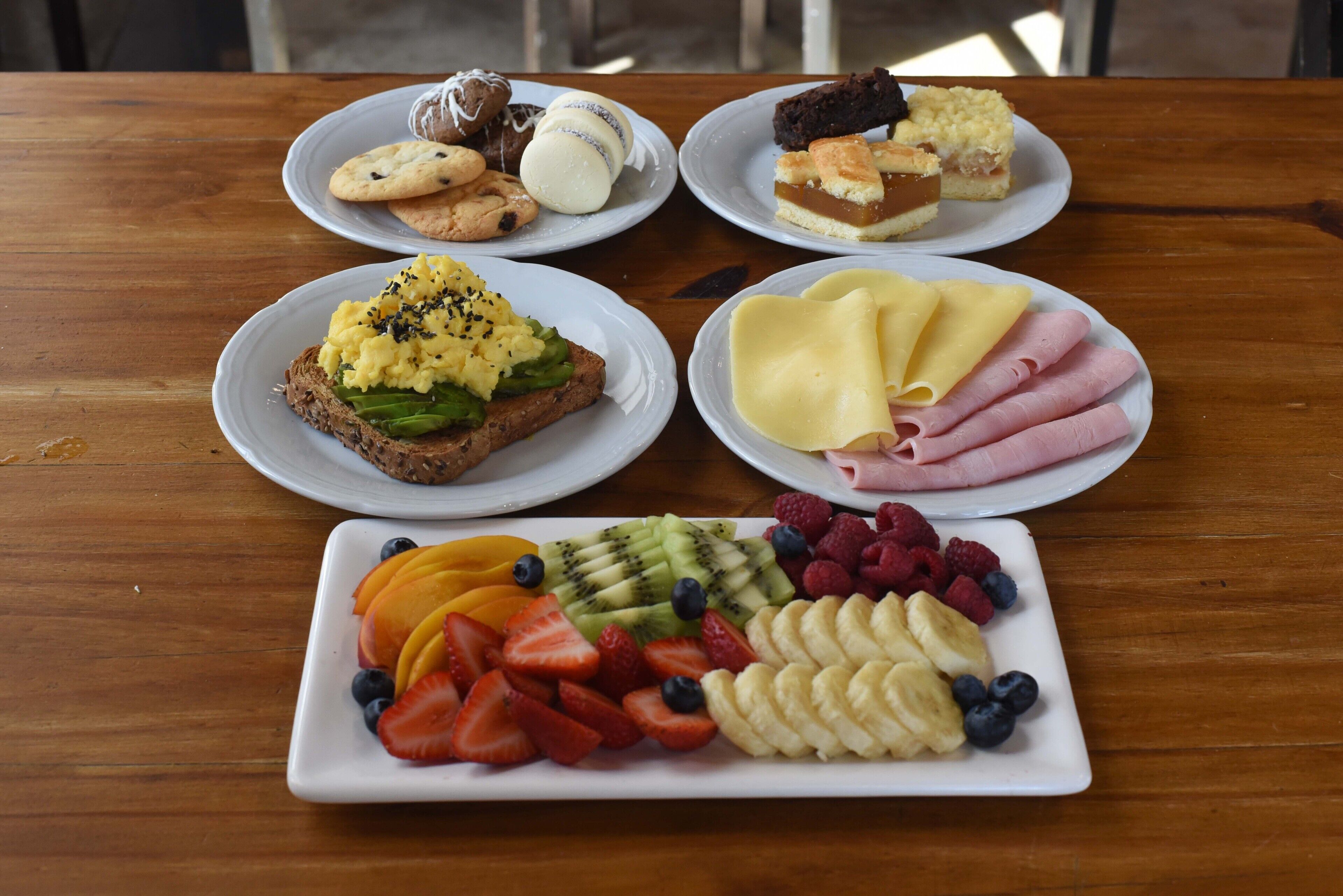 Una mesa de madera con diversos alimentos, incluyendo pasteles, sándwiches, queso, jamón y una bandeja de frutas.