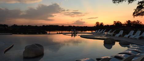 Un área de piscina tranquila con sillas de descanso y una gran roca como pieza central, rodeada de árboles y vistas al atardecer.