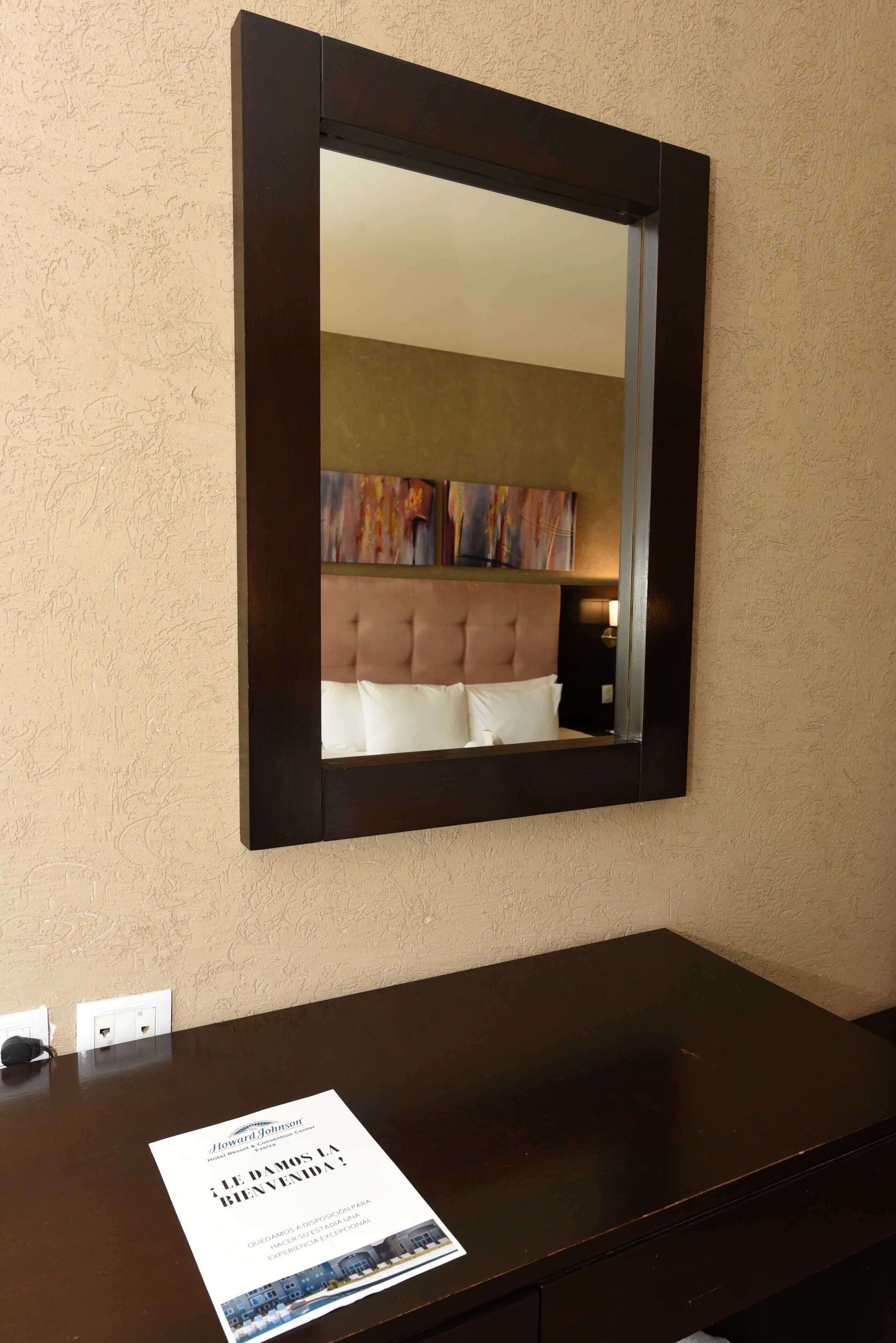 A hotel room with a large mirror, a dark wooden dresser, and a welcome brochure on the dresser.