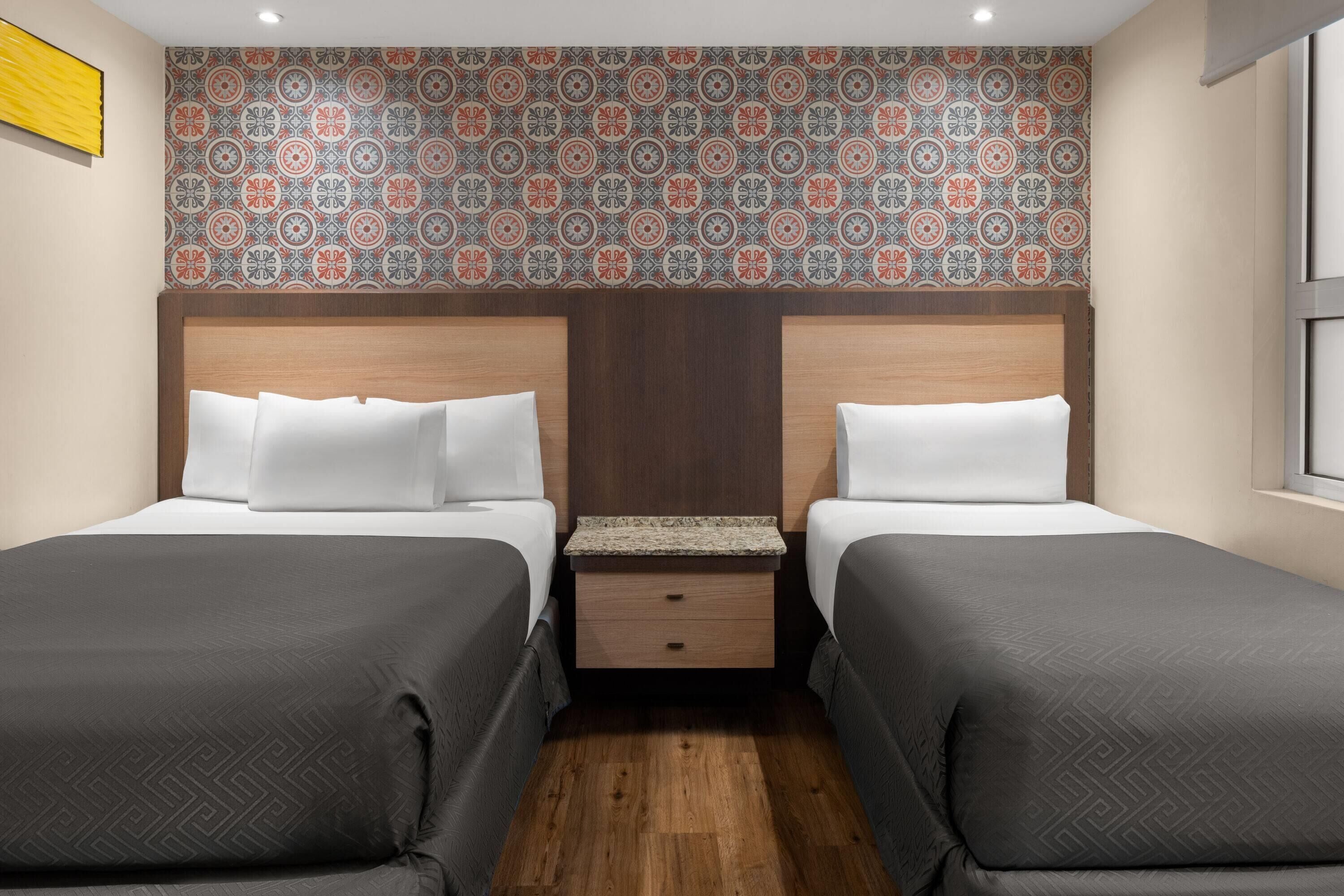 A hotel room with two beds, a wooden headboard, a small nightstand, and a patterned wallpaper.