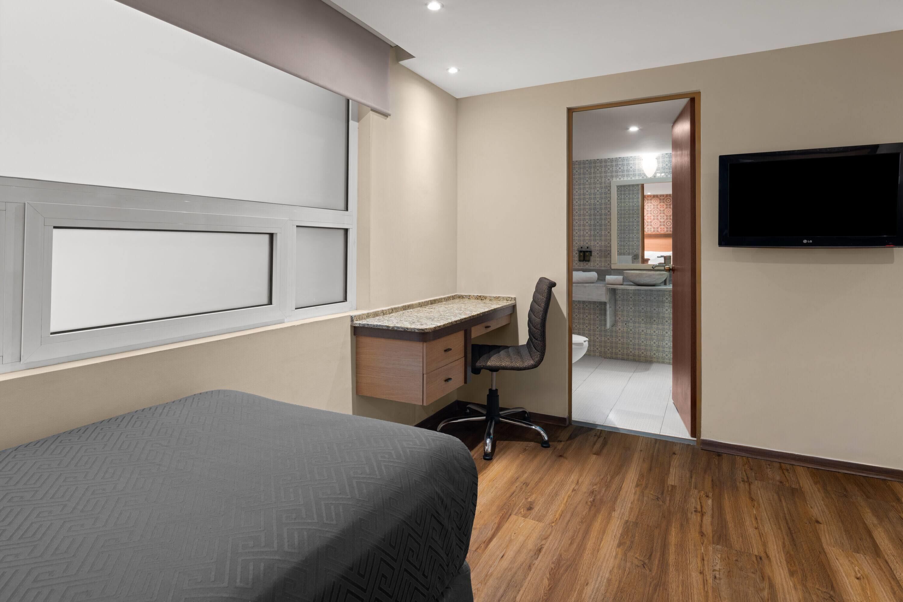 Un dormitorio con cama, un escritorio con silla, televisor y un baño visible a través de una puerta abierta.