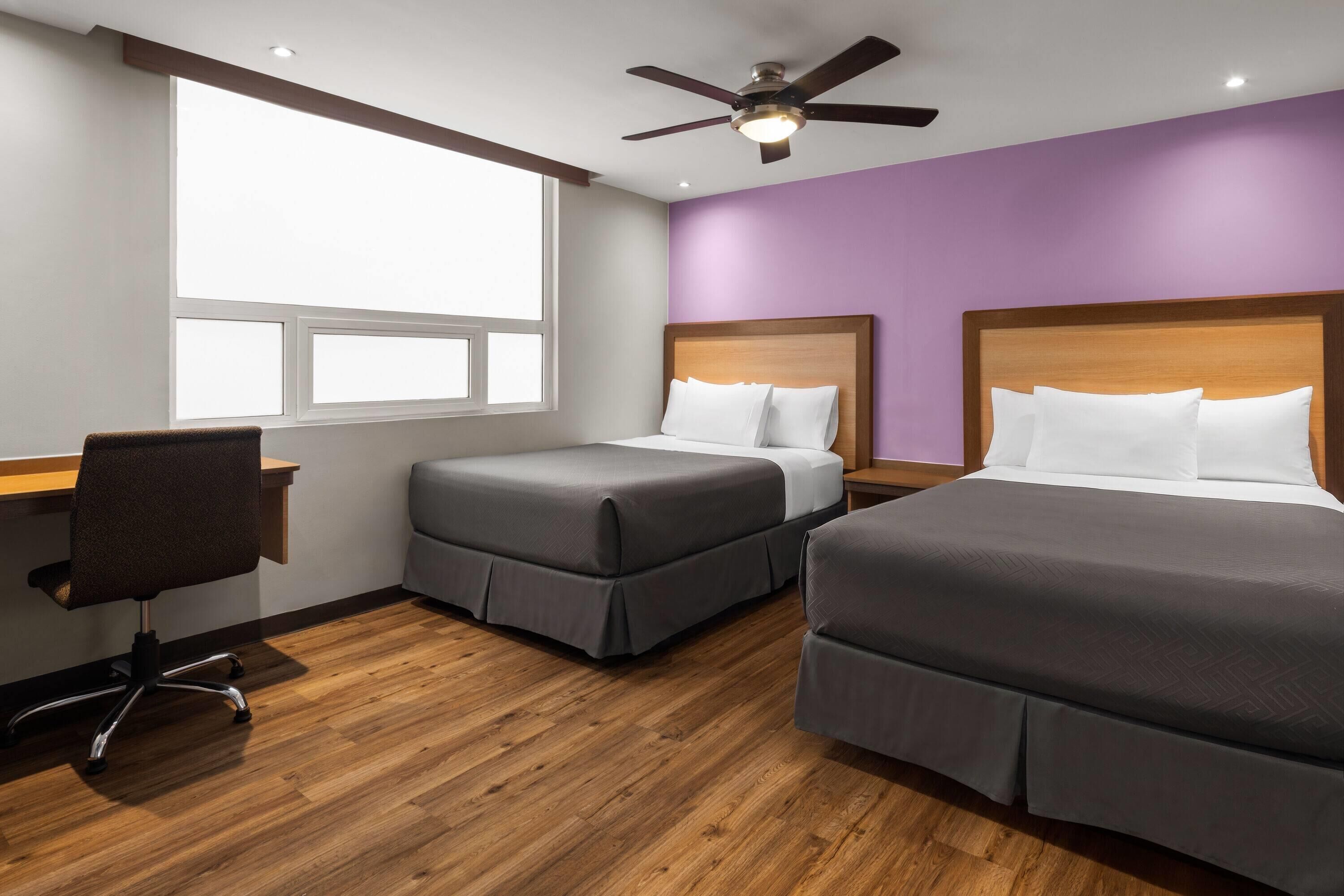 A hotel room with two beds, a desk, a chair, a ceiling fan, and a window providing natural light.