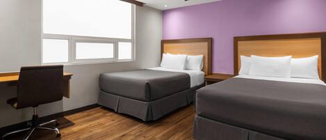 A hotel room with two beds, a desk, a chair, a ceiling fan, and a window providing natural light.