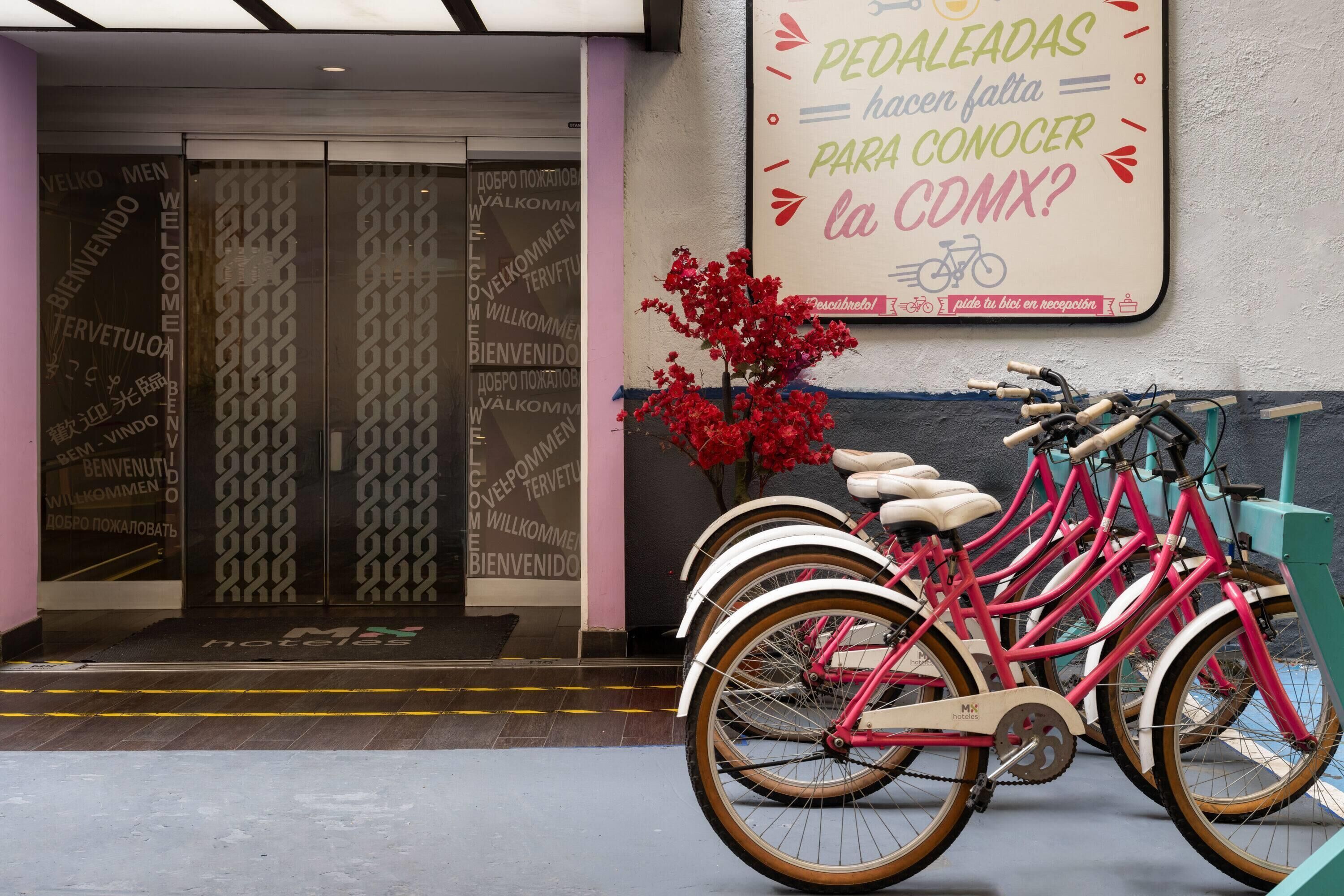 L’entrée d’un hôtel, avec une enseigne en langue étrangère, une composition florale et une rangée de vélos roses.