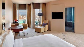 A hotel room with a large bed, a desk with a chair, a sofa, and a TV. There is a view of the city through the window.