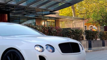 En vit Bentley stod parkerad framför ett hotell med en entré som bestod av ett glastak och en röd skylt.