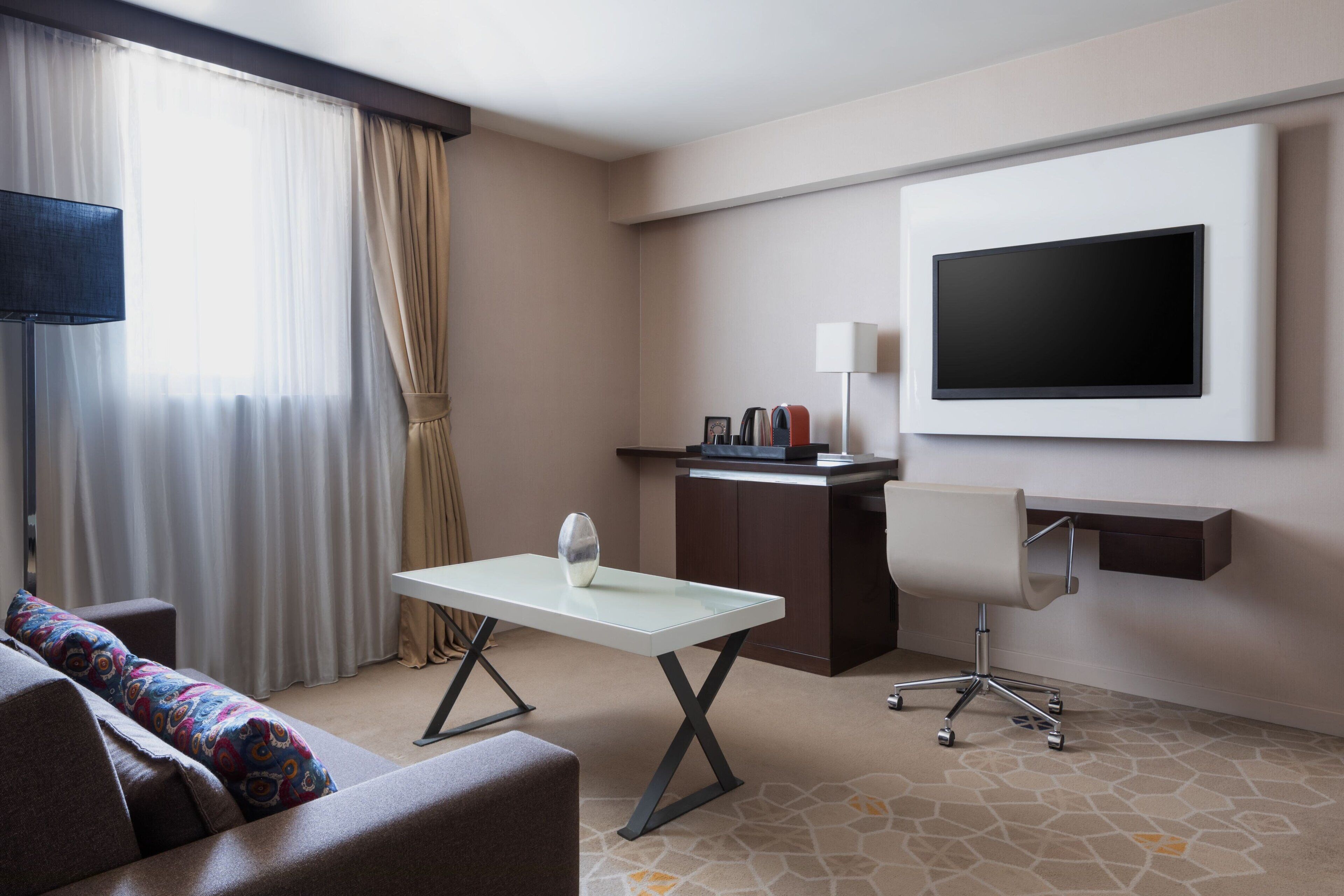 A hotel room with a sofa, a small table, a desk with a chair, a flat-screen TV mounted on the wall, and a window with curtains.