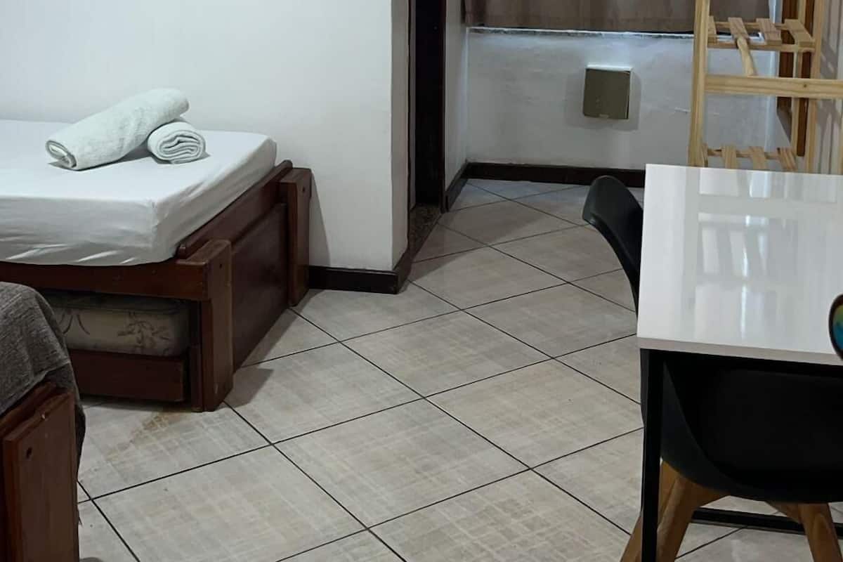 Ein kleines Hotelzimmer mit Bett, Tisch, Stuhl, einer Hängerträger aus Holz und einem Fenster mit Vorhängen.