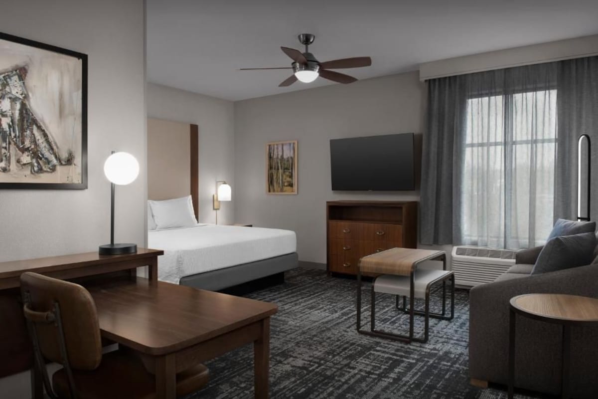 Une chambre d’hôtel équipée d’un lit, d’un bureau, d’une chaise, d’un canapé, d’une télévision, d’un ventilateur de plafond et d’œuvres d’art.