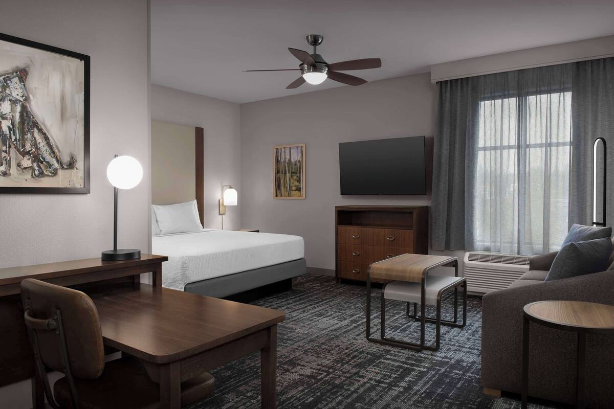 Ein Hotelzimmer mit Bett, Schreibtisch, Sessel, Sofa, Fernseher, Deckenventilator, Kunstwerken und einem Fenster mit Vorhängen.