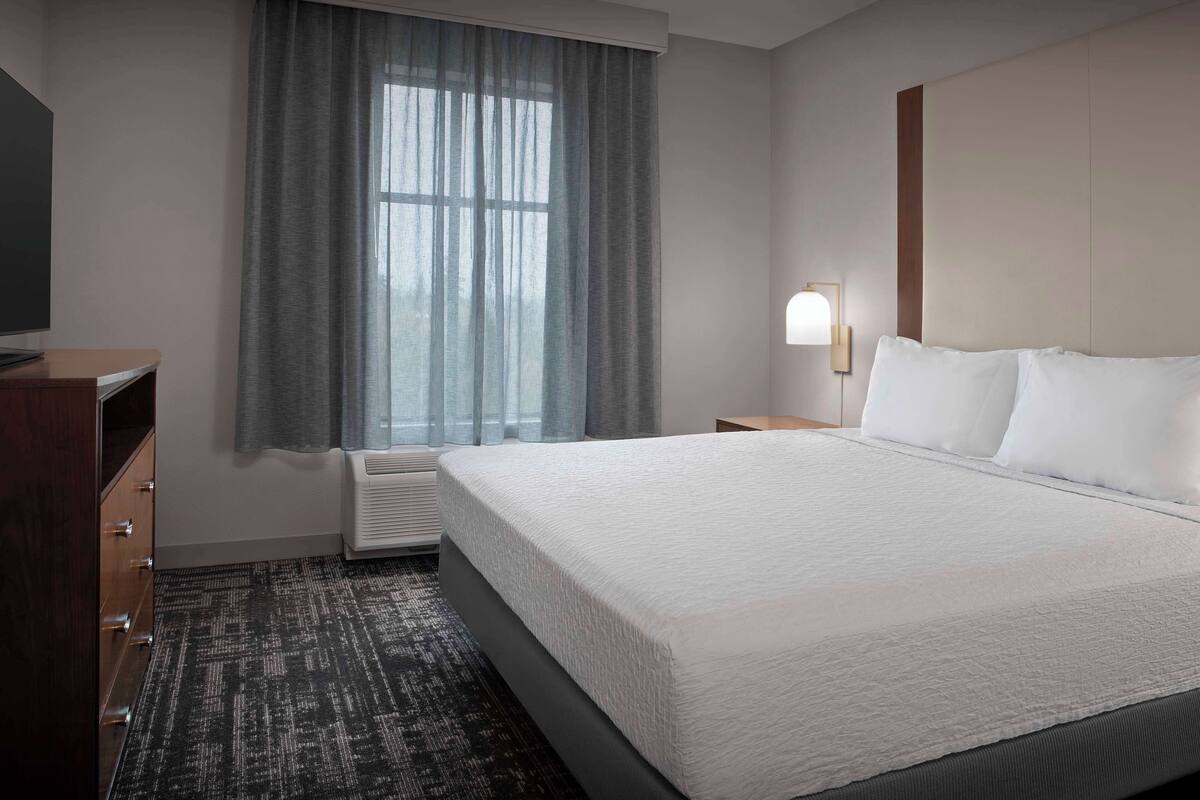 Ein Hotelzimmer mit einem großen Bett, einem Schreibtisch, einem Stuhl, einem Fernseher, einem Fenster mit Vorhängen und einer Wandlampe.