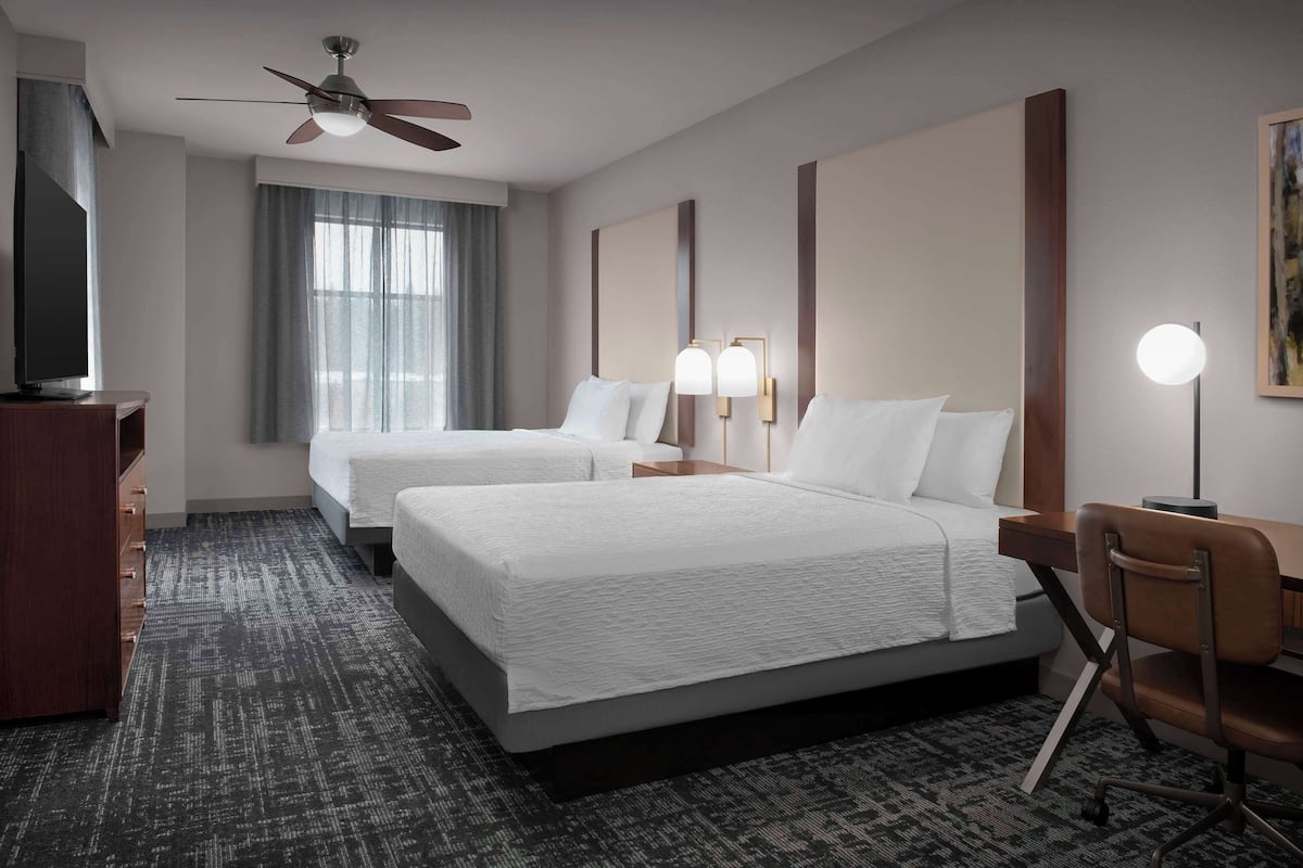 Ein Hotelzimmer mit zwei Betten, einem Schreibtisch, einem Sessel, einem Fernseher und einem Deckenventilator.