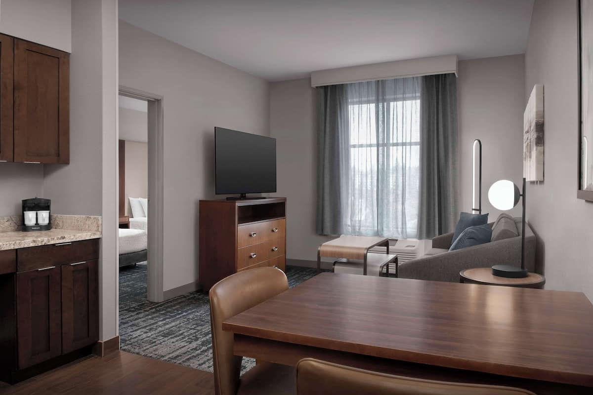 Ein modernes Hotelzimmer mit einem Holztisch, einem Flachbildfernseher, einer Couch und einem großen Fenster mit Vorhängen.