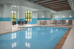 Una piscina interior con agua cristalina y azul, rodeada de sillas de descanso y mesas, y amplias ventanas que ofrecen luz natural.