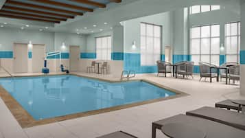 An indoor swimming pool with clear blue water, surrounded by lounge chairs and tables.