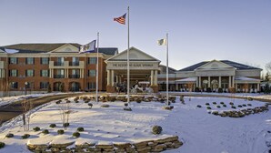 A hotel with a brick facade, a covered entrance, and a snow-covered landscape with stone planters and a flagpole. - The Resort at Glade Springs (Daniels)