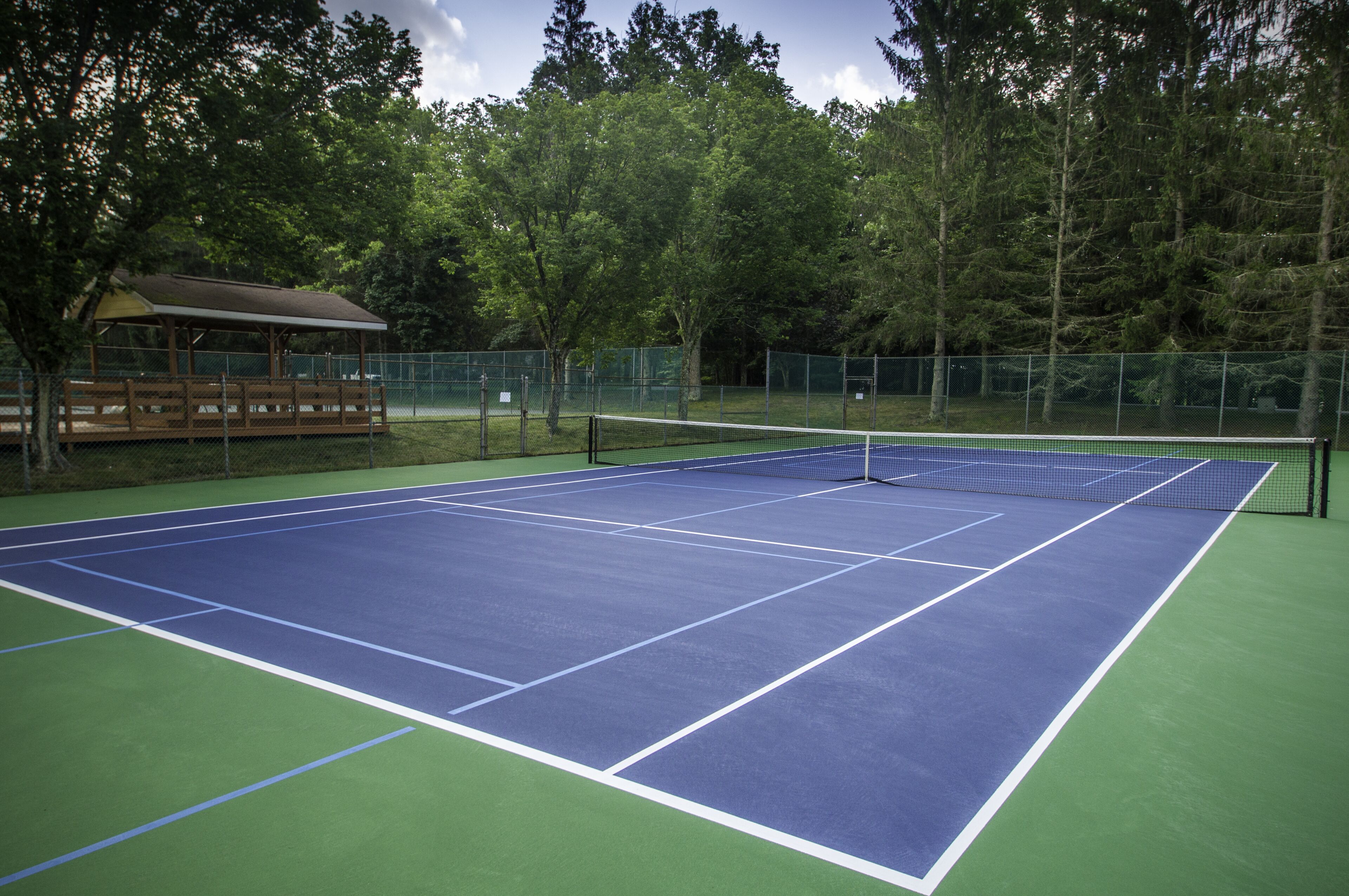 A tennis court with a net, surrounded by trees and a covered seating area.