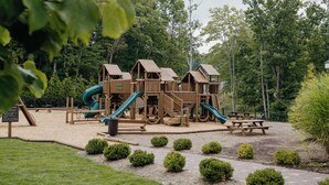 A wooden playground with slides and climbing structures, surrounded by trees and bushes. - The Resort at Glade Springs (Daniels)
