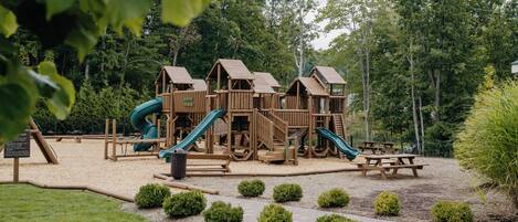 A wooden playground with slides and climbing structures, surrounded by trees and bushes.
