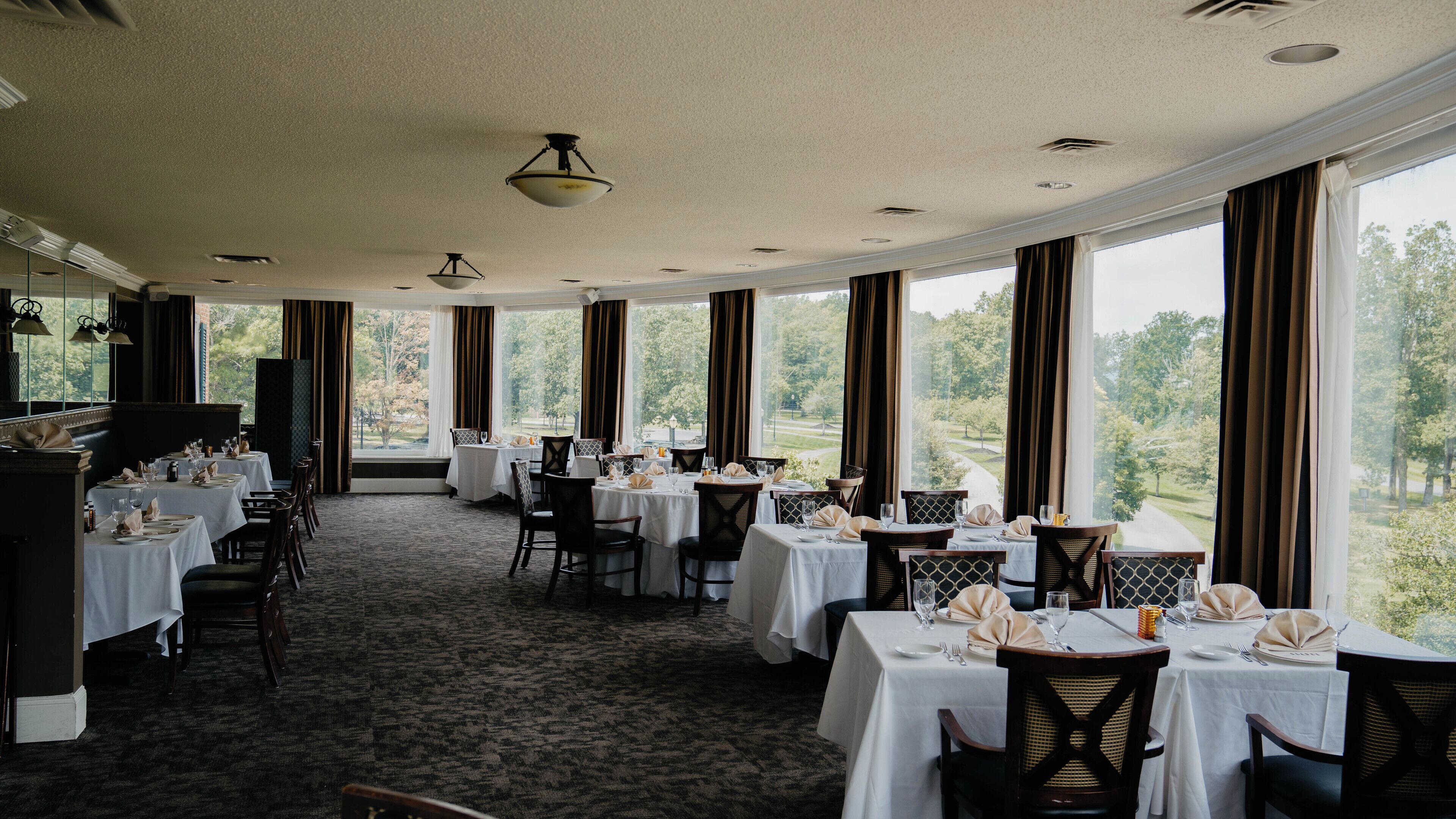 A dining area with tables set for service, large windows offering a view of the outdoors, and a carpeted floor.