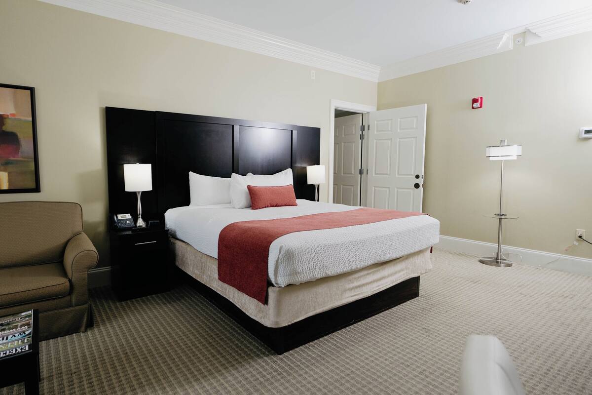 A hotel room with a bed, a nightstand, a chair, a painting, and a door.