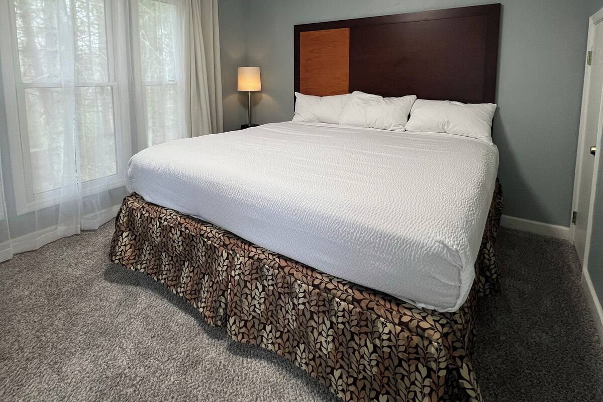 A neatly made bed with a patterned skirt, a bedside lamp, and a ceiling light in a room with light blue walls and a carpeted floor.