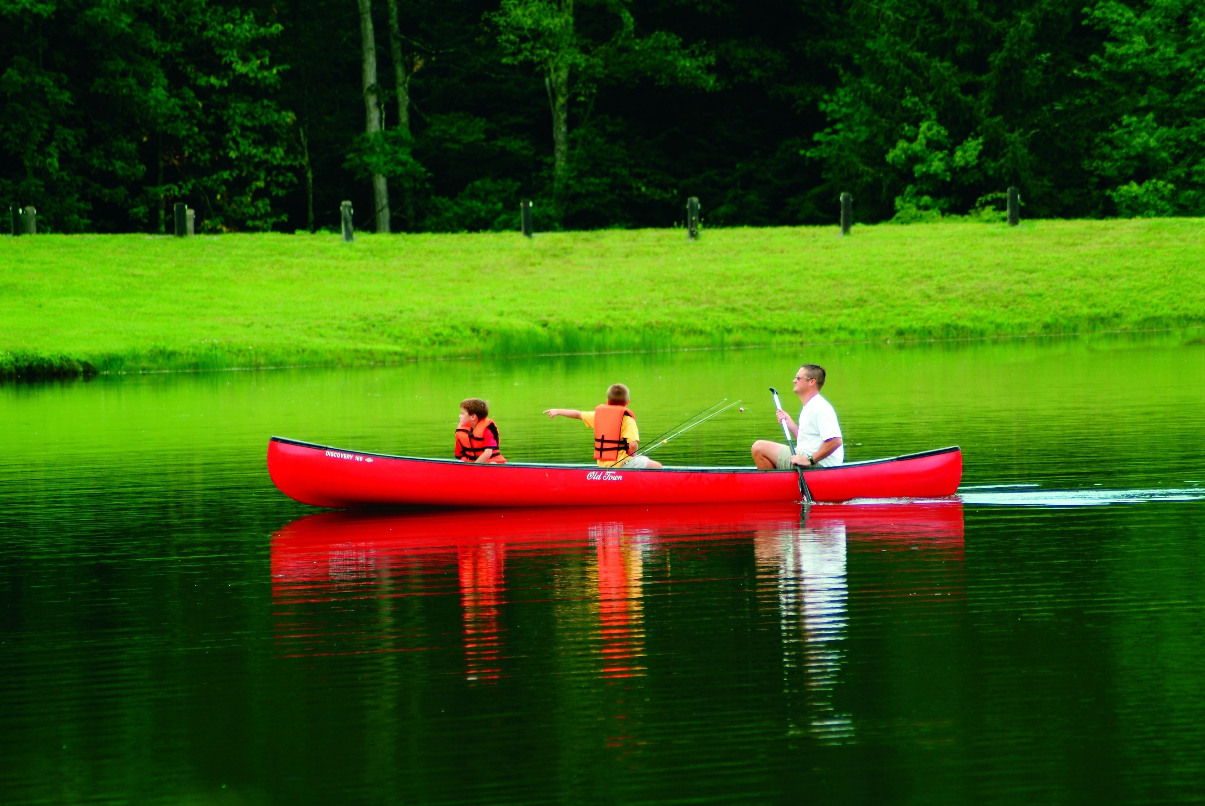 A red canoe on calm water with three people, surrounded by a grassy area and trees.