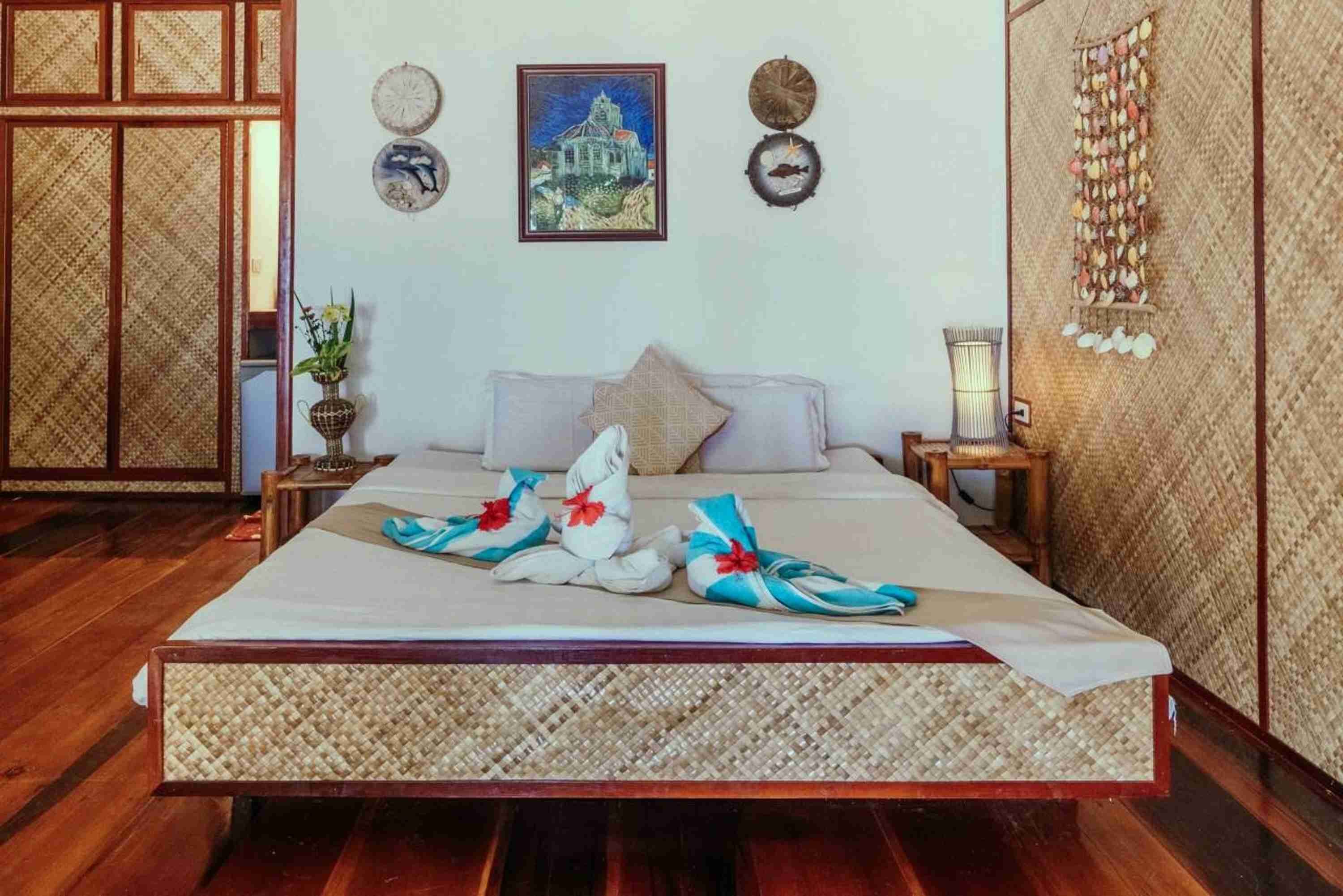 A neatly arranged bedroom with a large bed, a wooden floor, a traditional woven partition, and a wall decorated with cultural artifacts.