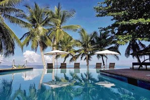Un área junto a la piscina con sillones reclinables y sombrillas, rodeada de palmeras y una vista despejada del océano.