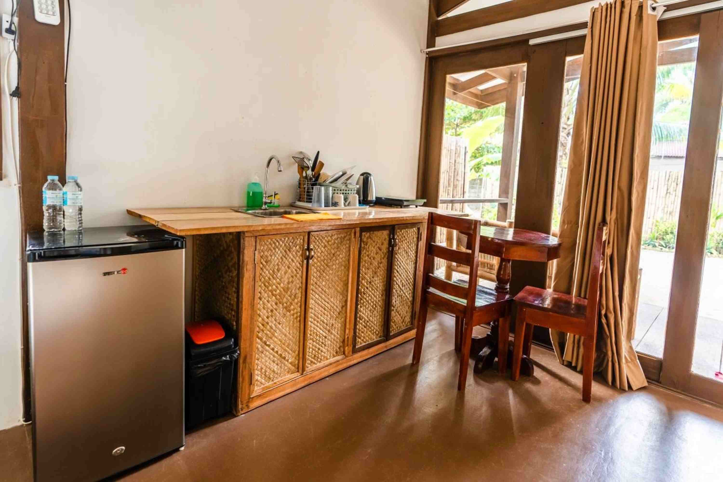 A compact kitchen with a refrigerator, sink, and wooden cabinet. There is a table and chair set near a window with curtains.