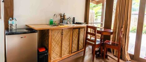 A compact kitchen with a refrigerator, sink, and wooden cabinet. There is a table and chair set near a window with curtains.