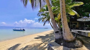 Una playa con un bote, palmeras y un área techada con mesas y sombrillas.
