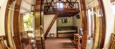 Una habitación acogedora con cama, una mesita y una silla. Hay una ventana con cortinas, techo de madera y una escalera que sube a un nivel superior.