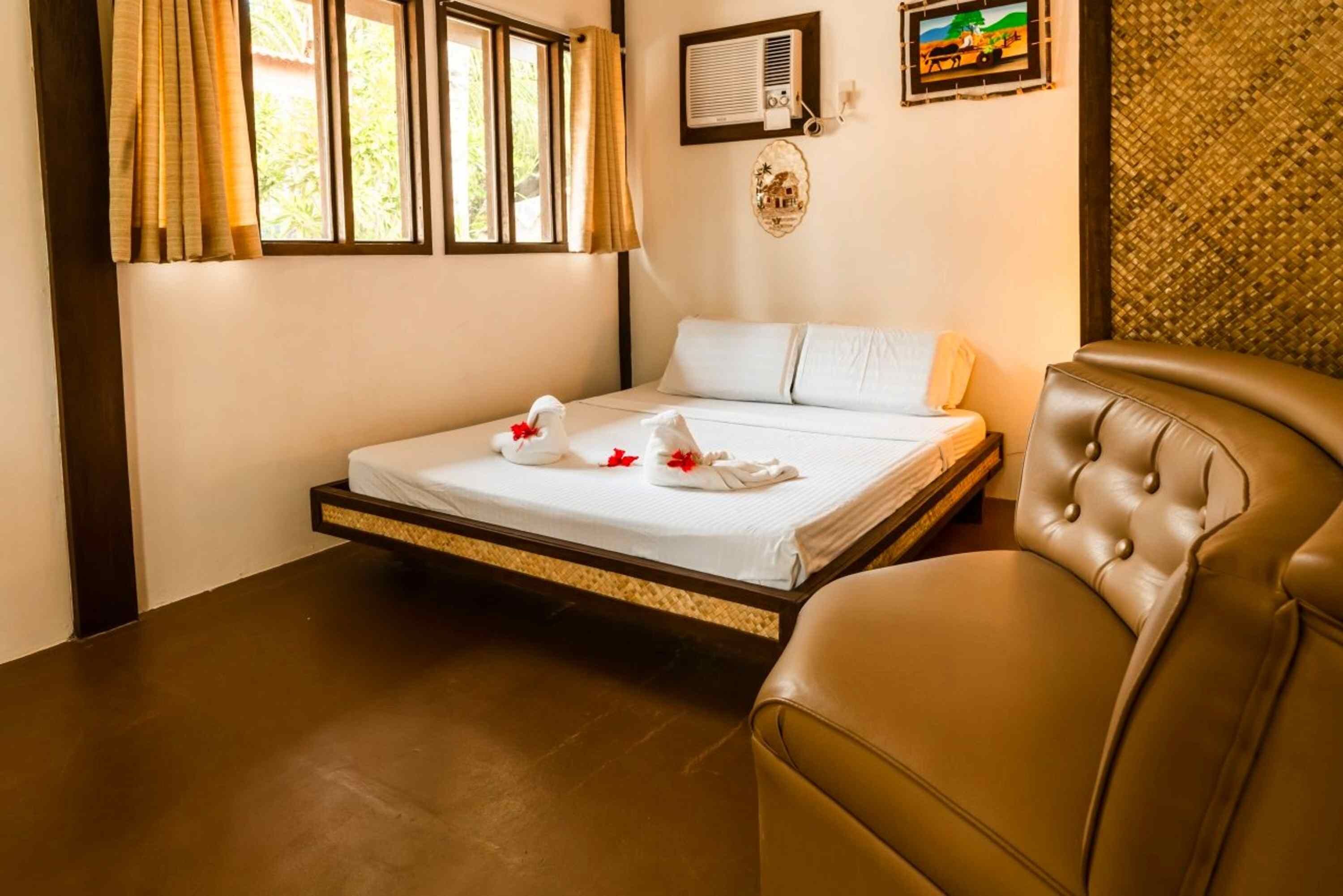A hotel room with a bed, a brown leather chair, a window with curtains, and a wall-mounted air conditioner.