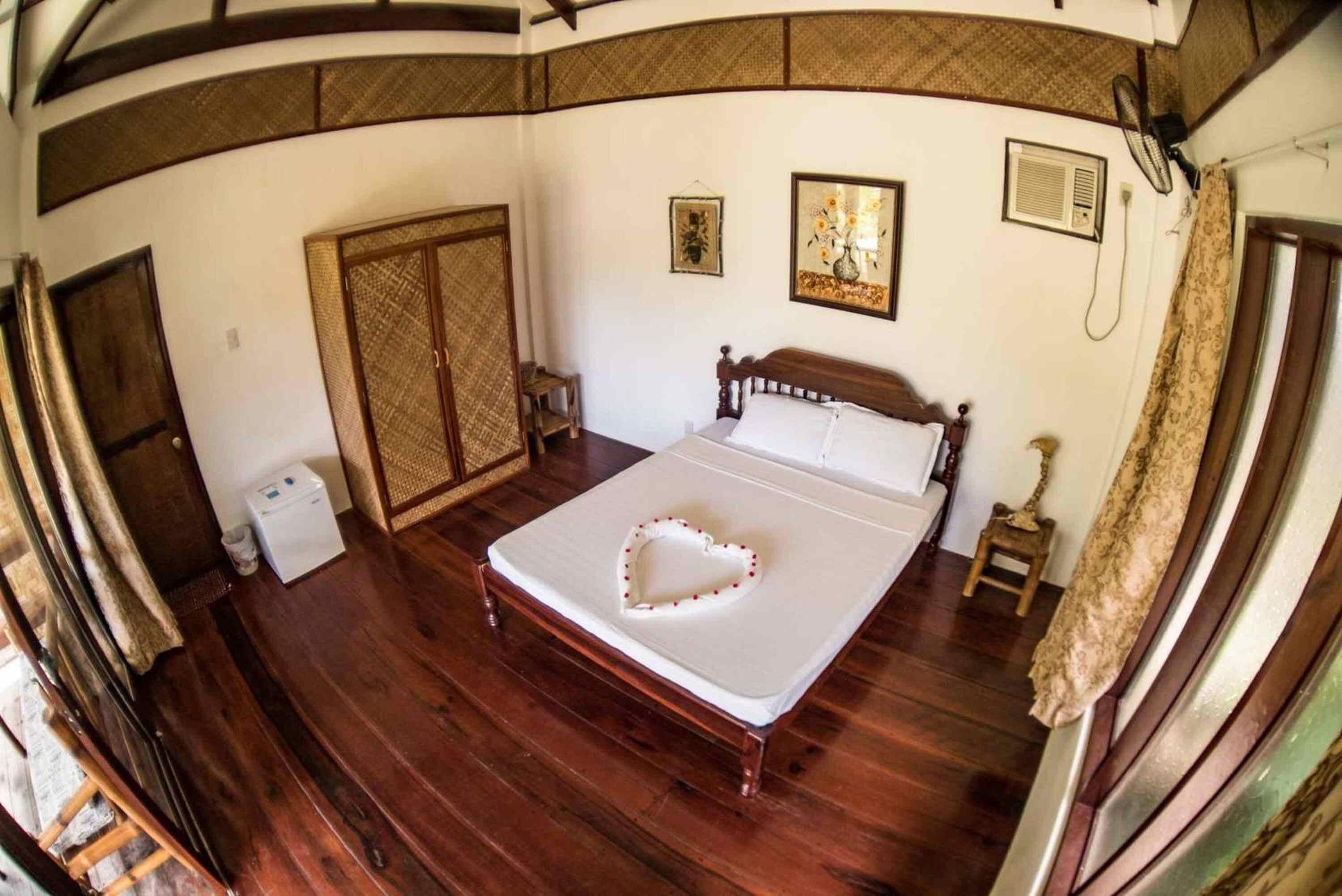 Een kamer met een houten bed, een kledingkast, een kleine tafel en een ventilator.