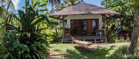 A thatched-roof hut with a porch, surrounded by lush tropical vegetation.