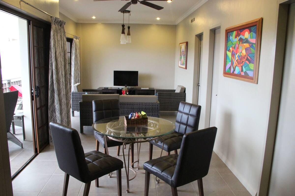 A modern living room with a glass-top dining table surrounded by leather chairs, a flat-screen TV mounted on the wall, and a colorful abstract painting.