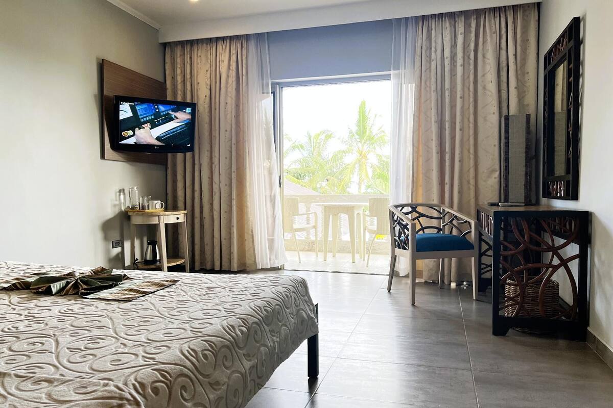A hotel room with a bed, a desk, a chair, a television, and a balcony with a view of palm trees.